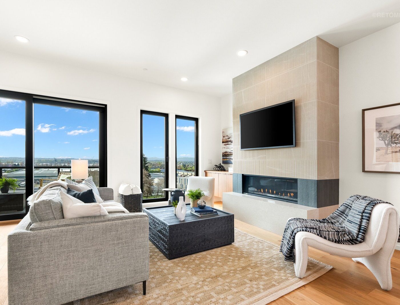 Modern living room with a grey sectional, a fireplace with a mounted TV, and large windows showing a city view.