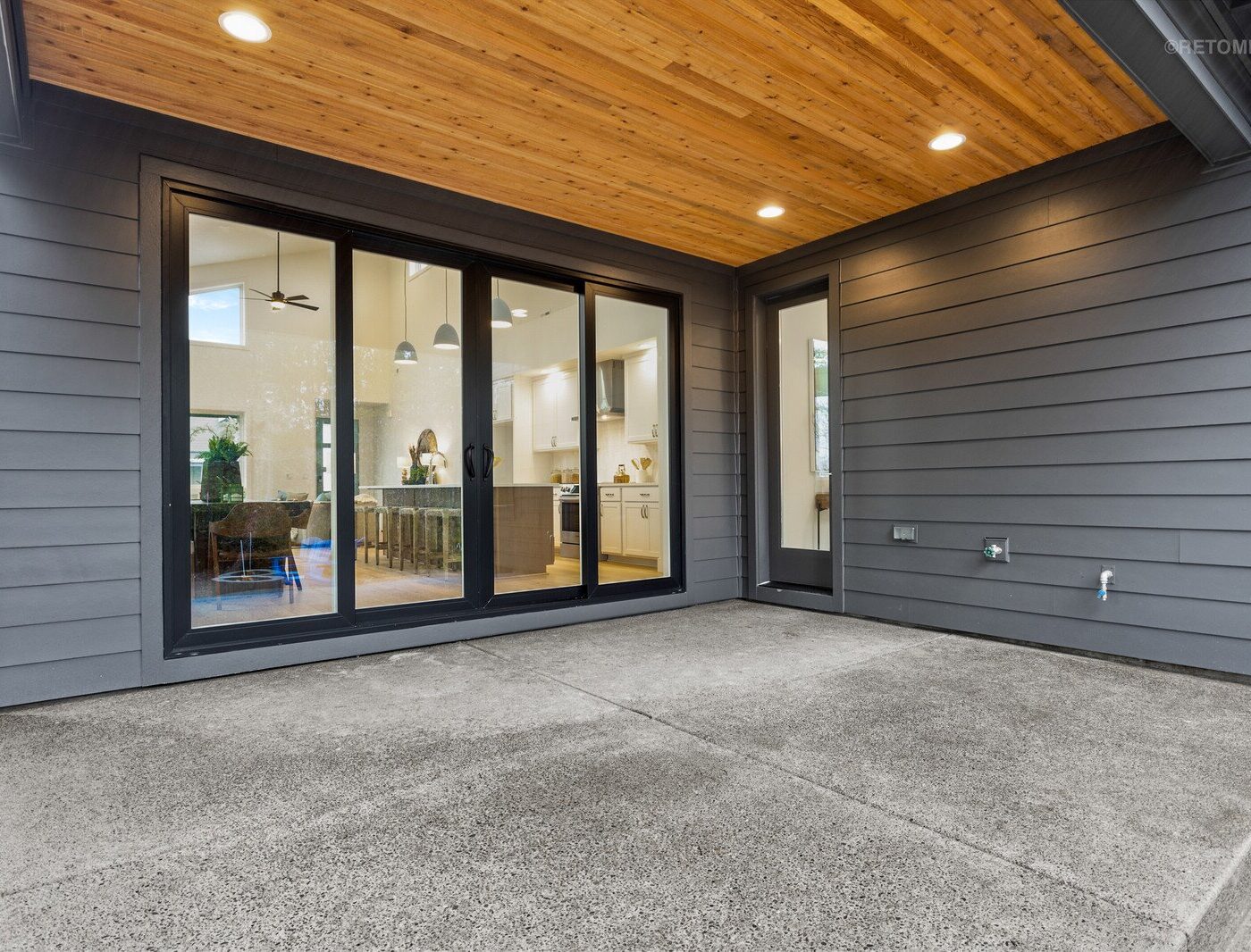Concrete patio area under a wood-paneled ceiling with sliding glass doors leading into a modern kitchen.