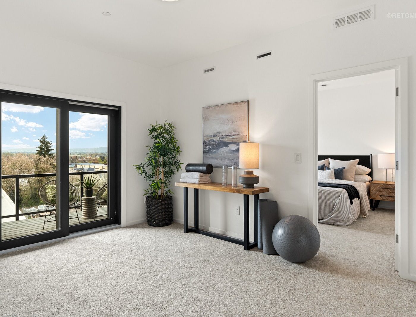 Bright modern room with light gray carpet, a desk area with a lamp, and sliding glass doors leading to a balcony.
