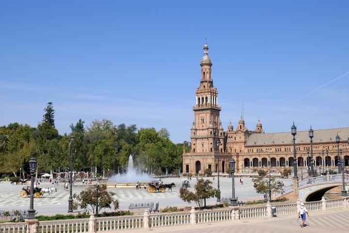 Plaza de España in Seville with its tall ornate tower, a fountain, and horse-drawn carriages under a clear blue sky.