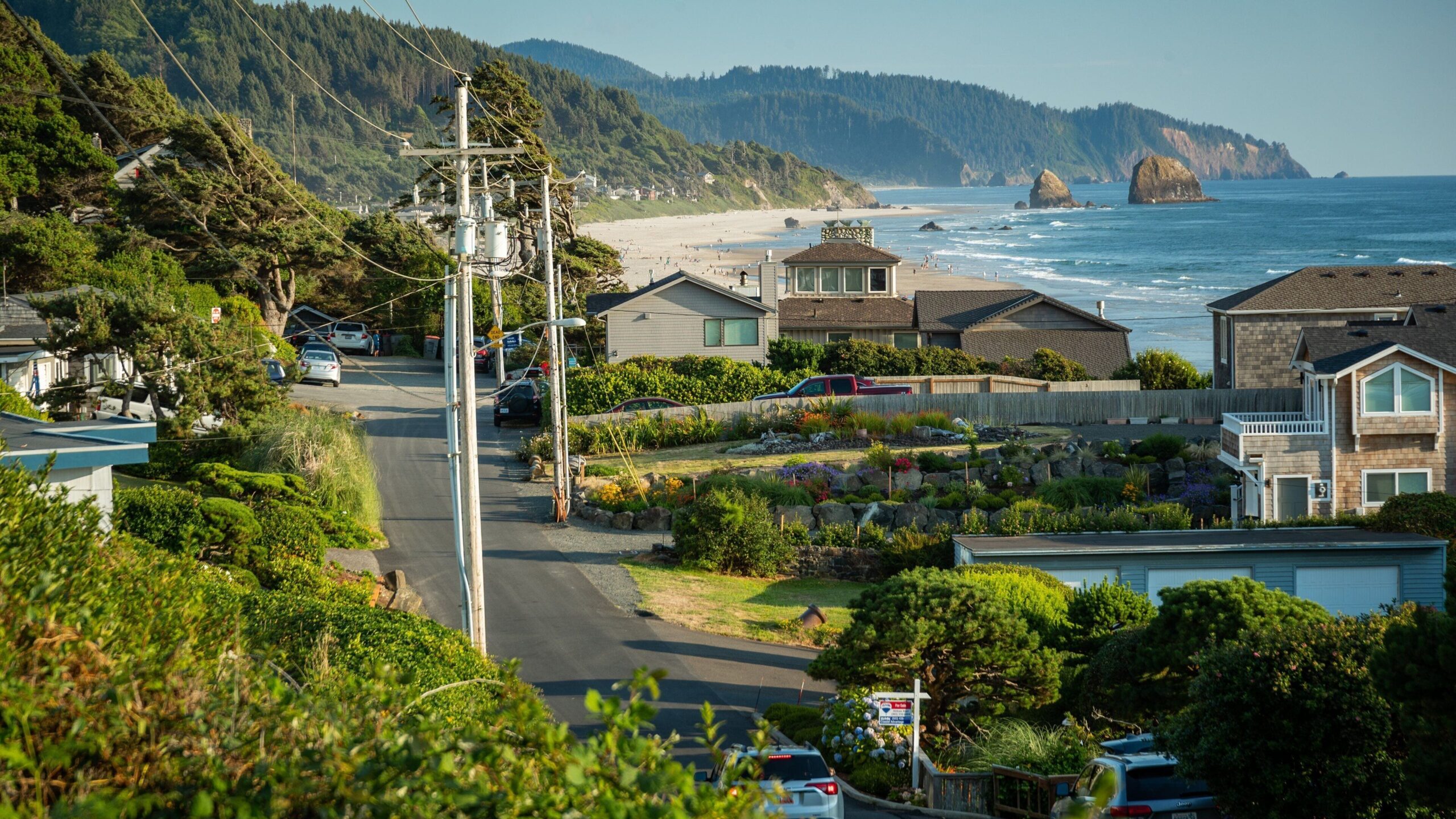 Coastal road leading toward a beach with houses, forested hills, and sea stacks under a clear sky.