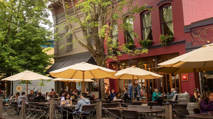 People sitting at outdoor cafe tables under beige umbrellas in front of red and grey buildings.