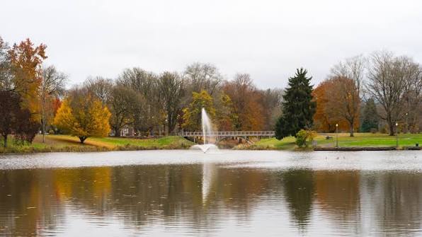 A water fountain sprays upward in a calm pond, surrounded by autumn trees in shades of gold, orange, and green.