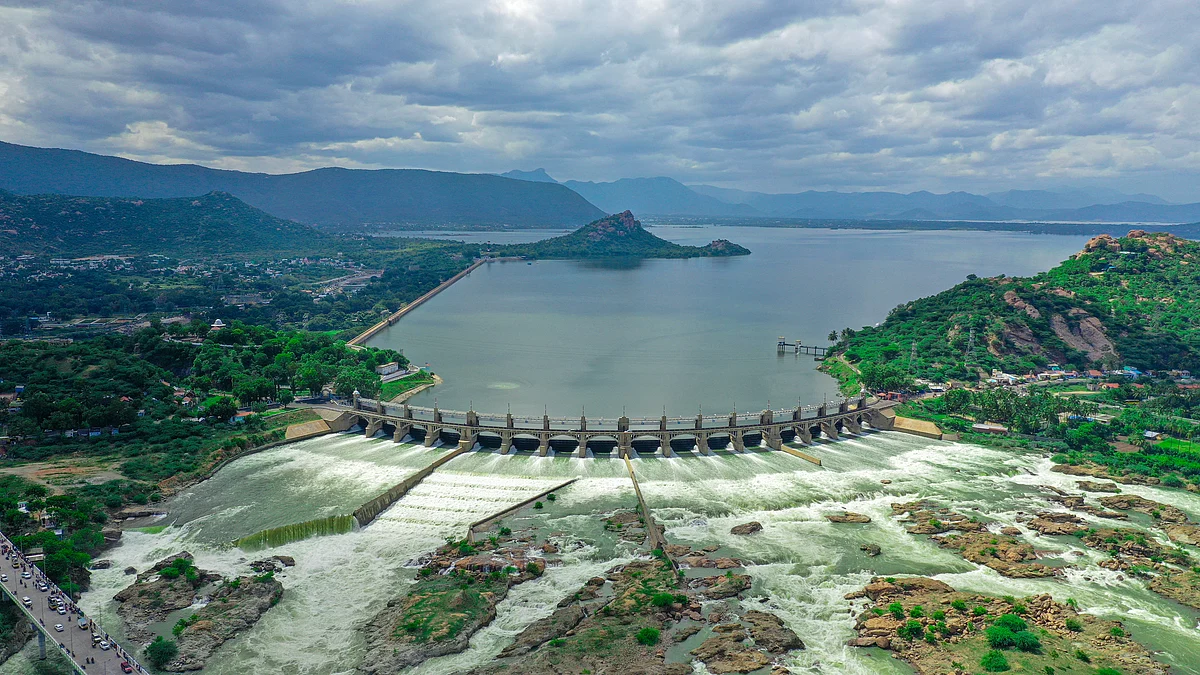 Aerial view of a curved dam with rushing white water flowing downstream toward rocky terrain under a cloudy sky.